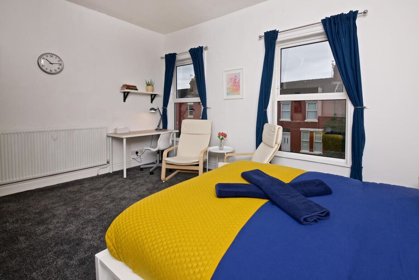 A hotel room with a bed, two chairs, a desk, a clock, and a window with curtains.