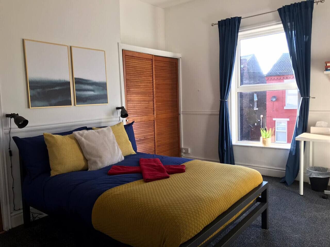 Un dormitorio con cama, ventana con cortinas, escritorio y vista a un edificio rojo en el exterior.