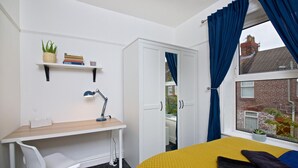 Un dormitorio con cama, escritorio, silla, ventana con cortinas y vista a un edificio de ladrillo.