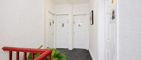 A hallway with white textured walls, a red railing, and a plant. There are two white doors with a small sign above each.