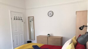 A bedroom with a bed, a bedside table, a mirror, a clock, and a door with coat hooks. - Experience Comfort in Liverpool (Merseyside)