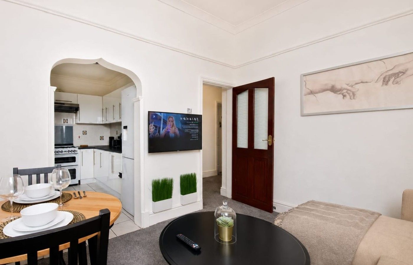 Una sala moderna con comedor, una cocina al fondo, un televisor de pantalla plana montado en la pared y una puerta que da a otra habitación.