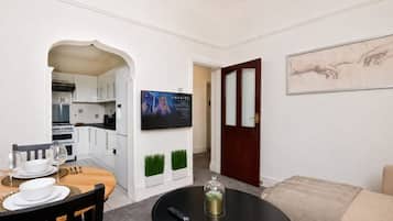 Una sala moderna con comedor, una cocina al fondo, un televisor de pantalla plana montado en la pared y una puerta que da a otra habitación.