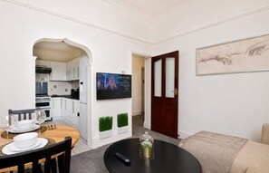 A modern living room with a dining area, a kitchen in the background, a flat-screen TV mounted on the wall, and a door leading to another room. - Experience Comfort in Liverpool (Merseyside)