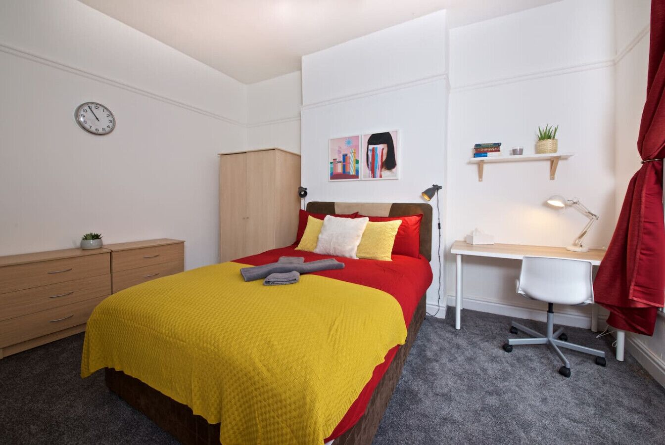 A bedroom with a bed, a desk, a chair, a wardrobe, and a clock on the wall.