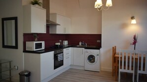 Een kleine keuken met een magnetron, oven en wasmachine, een zithoekje met een tafel en stoelen, en een hangende lamp.