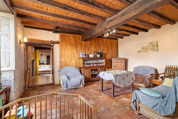 A rustic room with wooden beams, a dining table, chairs, a bookshelf, and a crib.