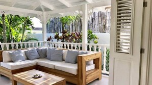 Un espace extérieur aménagé avec un coin salon abrité, comprenant un canapé en bois et une table basse, le tout entouré d’une rambarde blanche et de plantes.