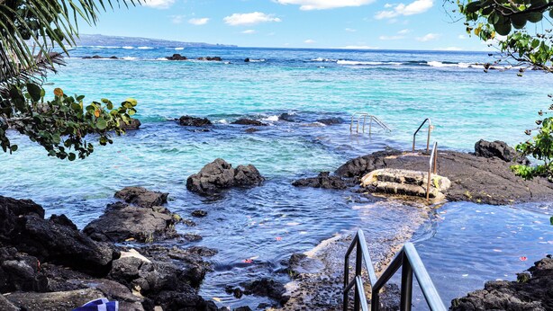 A rocky shoreline with clear turquoise water, a metal staircase leading into the sea, and a distant view of a coastline with buildings. - Sea Turtle Suite - Steps from Carlsmith Beach! (Hilo)