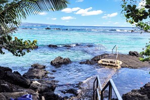 A rocky shoreline with clear turquoise water, a metal staircase leading into the sea, and a distant view of a coastline with buildings.