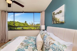 A bedroom with a large window, a ceiling fan, a bed with floral bedding, a bedside lamp, and a landscape painting on the wall.