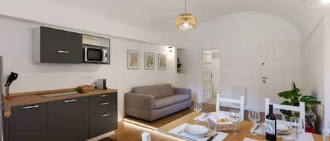 Une cuisine moderne équipée d’un four à micro-ondes encastré, un coin repas avec une table dressée pour deux personnes, et un canapé en arrière-plan.