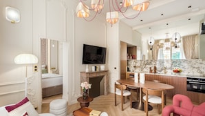 A modern living room with a sofa, a coffee table, a TV, and a fireplace. There is a dining area with a wooden table and chairs. The kitchen is visible in the background with a marble countertop and modern appliances. - Merveil Signature Suite - Marignan (Paris)