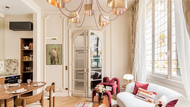 A well-lit living room with a dining area, a sofa, and a red armchair. There is a wooden coffee table with books and a vase of flowers. - Merveil Signature Suite - Marignan (Paris)