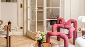 A room with a wooden floor, a pink chair, a wooden coffee table, and a white lamp. There is a door with glass panels leading to a closet. - Merveil Signature Suite - Marignan (Paris)