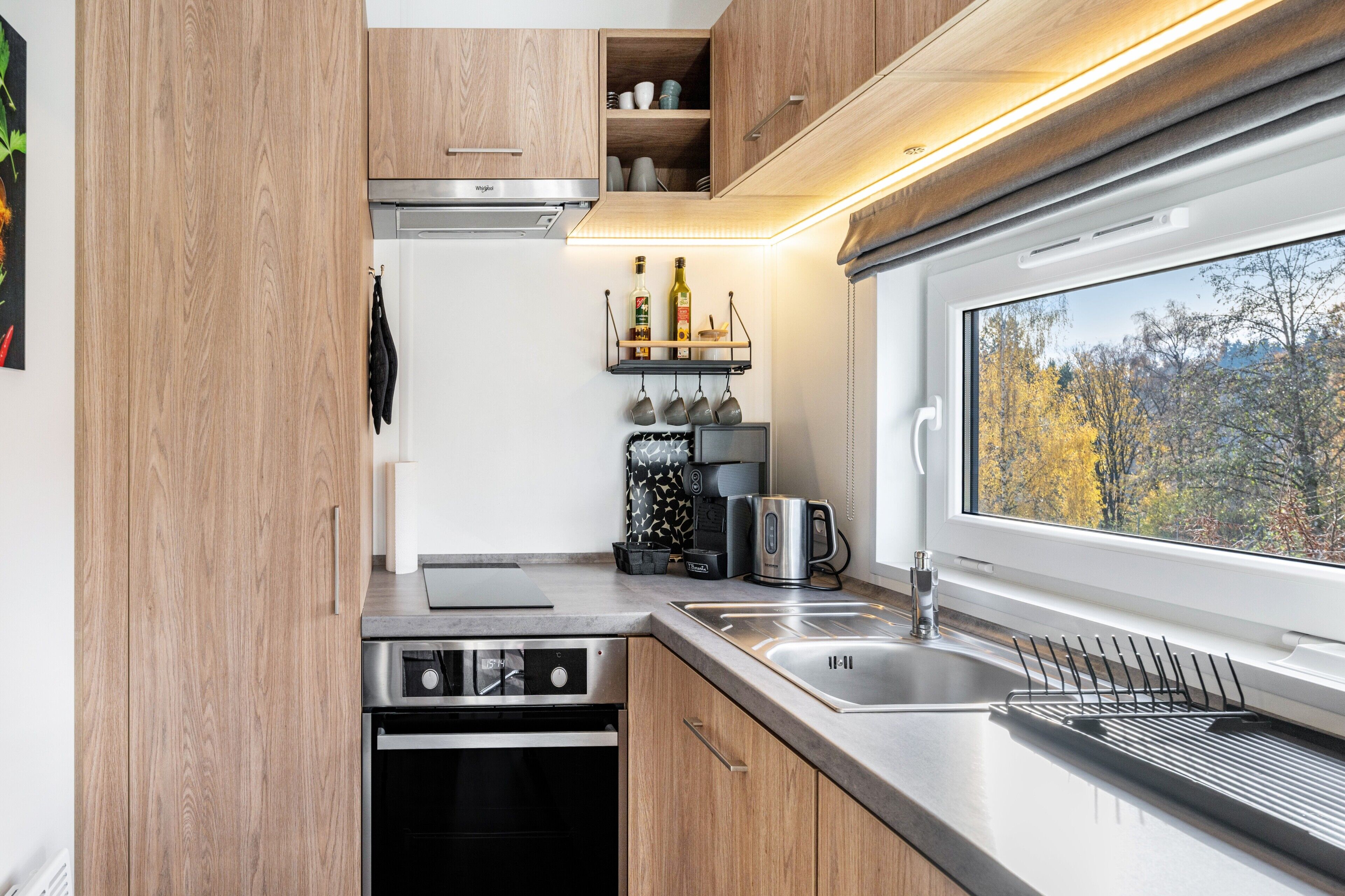 A compact kitchen with wooden cabinets, a built-in oven, a sink, and a window with a view of trees.