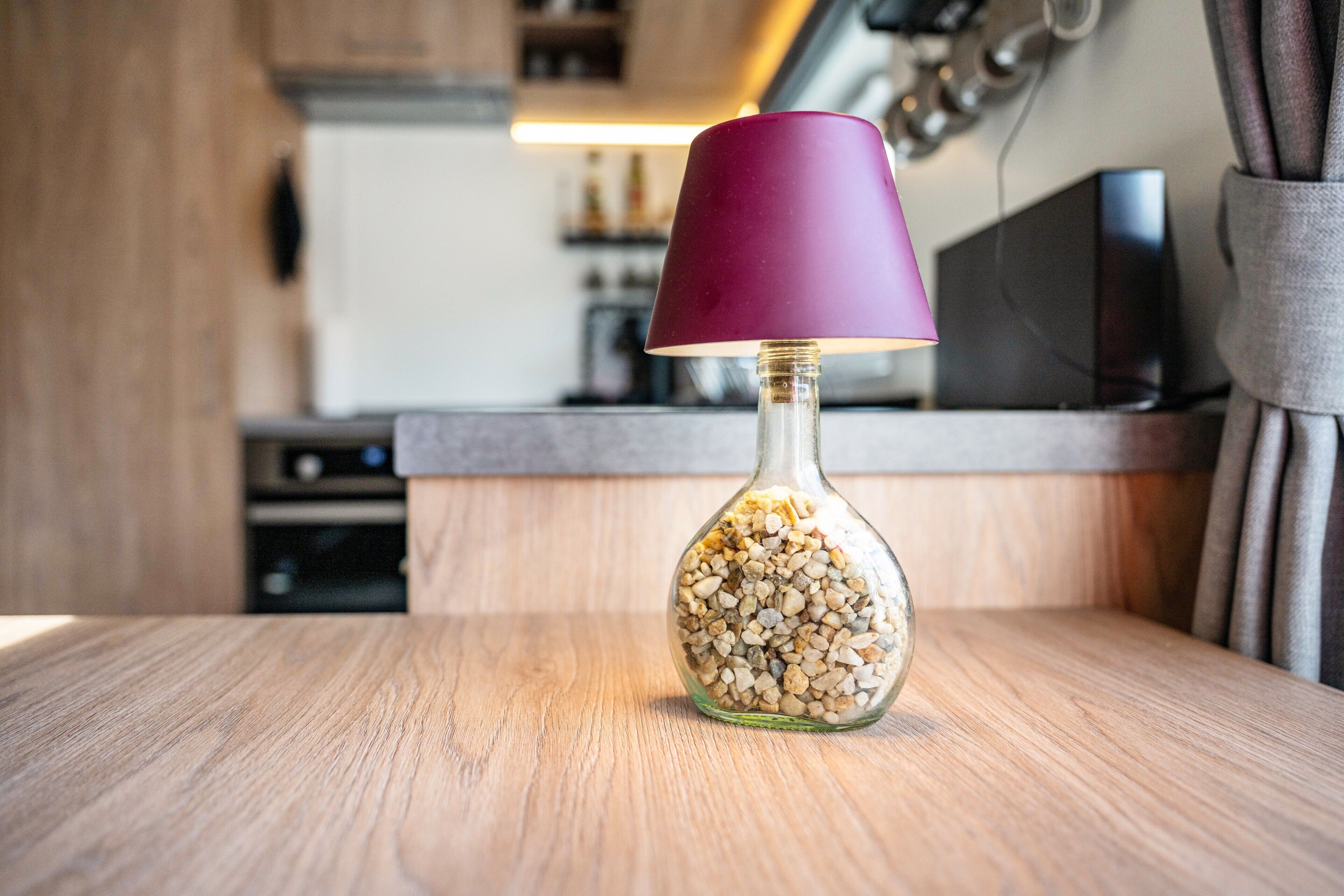 A glass bottle lamp with a purple shade on a wooden surface in a kitchen.