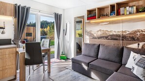 A modern living room with a sofa, a dining area with a table and chairs, a bookshelf, and a view of the outdoors.