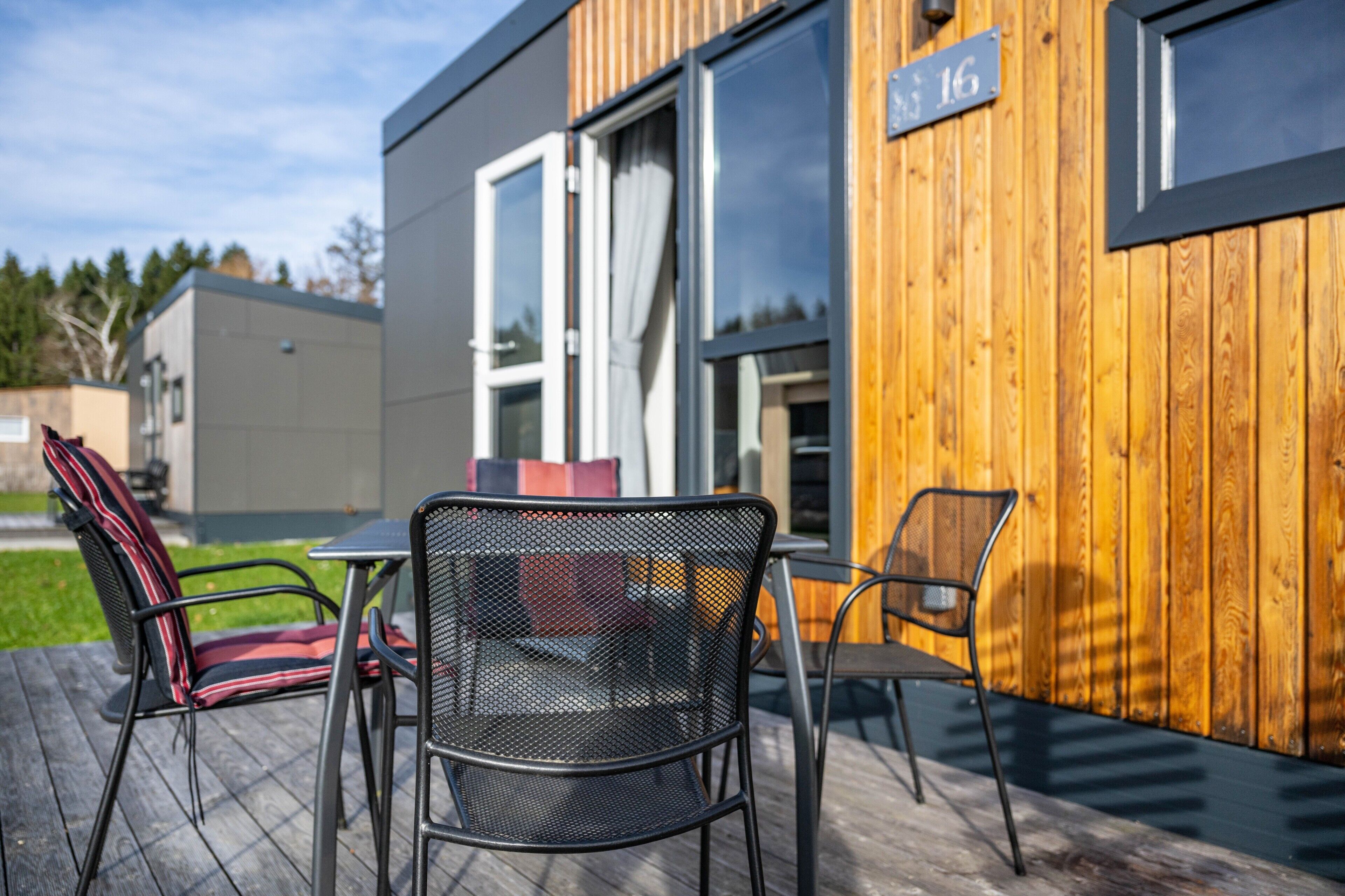 Un bâtiment moderne avec une façade en bois, une enseigne portant le chiffre « 16 », et un espace extérieur aménagé avec des chaises et une table en métal.