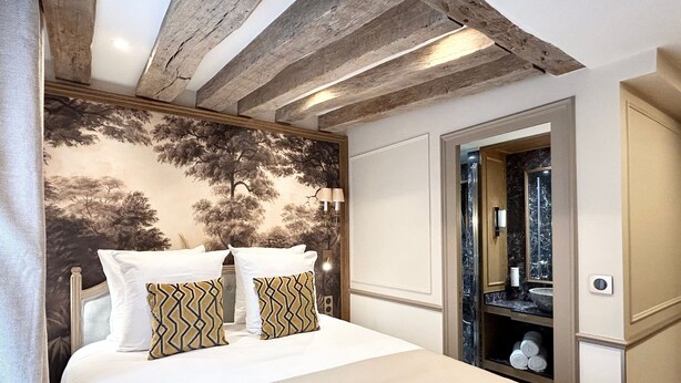 Quarto com cama, duas almofadas, cabeceira com mural de árvore, teto de madeira e banheiro visível por uma porta aberta.