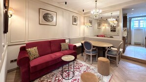 Uma sala de jantar com um sofá vermelho, uma mesa redonda e cadeiras. Há molduras de quadros nas paredes e um lustre.