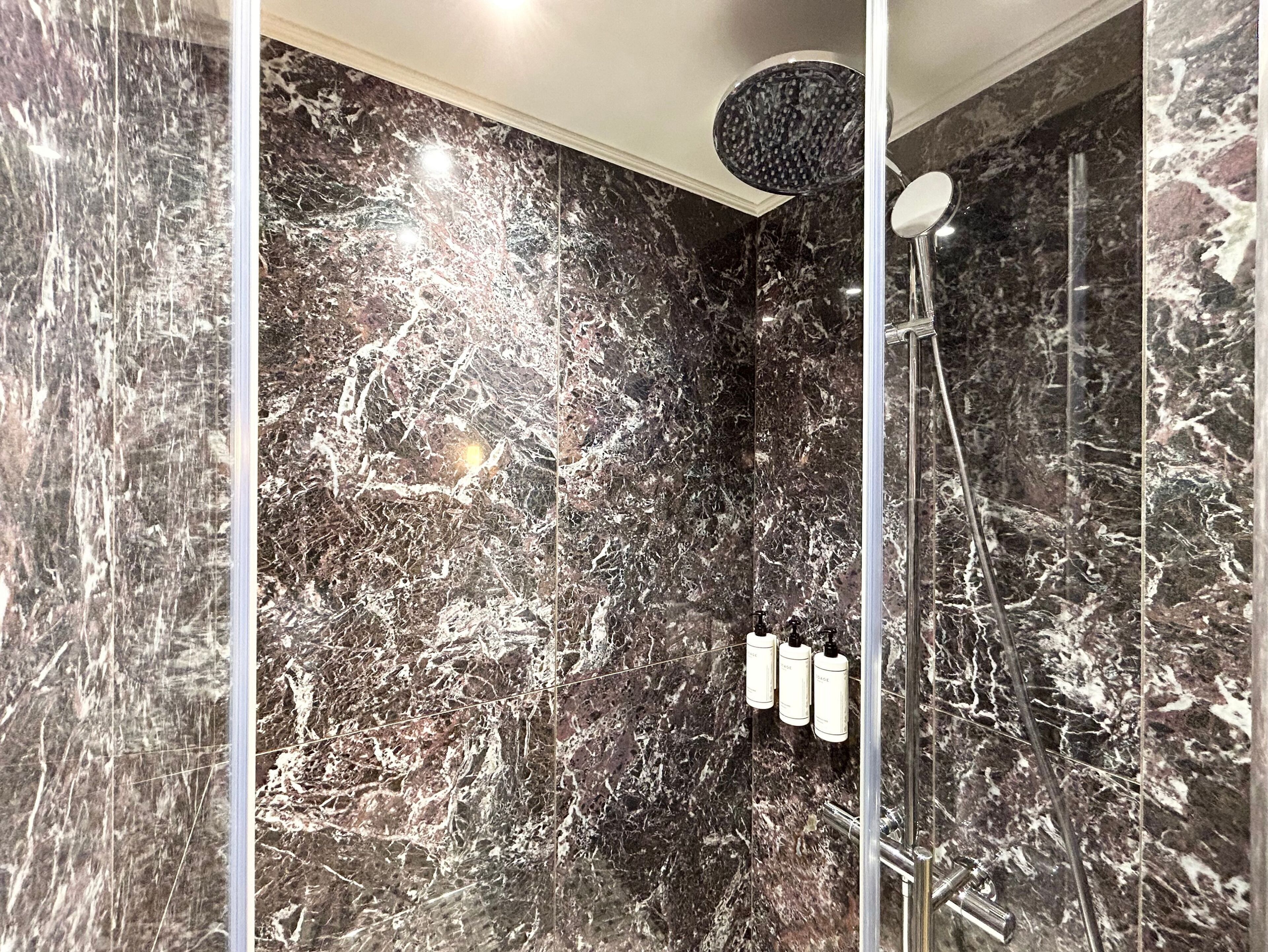 A modern bathroom with a marble wall, a shower with a round head, and three bottles of shampoo.