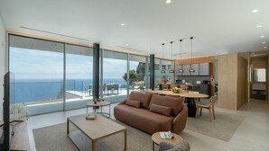 A modern living room with a large window overlooking the sea, a dining area, and a kitchen. - Superb Top Luxury Villa|GrandView Cristo Rei (Canico)