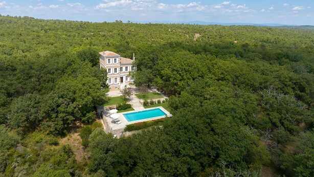 A large, multi-story building surrounded by dense forest, with a swimming pool and outdoor seating area. - House on large 200-hectare estate with swimming pool (Esparron)