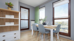 Ein Essbereich mit Holztisch, vier grauen Stühlen, einer weißen Kommode mit Schubladen, einer Topfpflanze auf einem Regal und großen Fenstern mit Vorhängen.