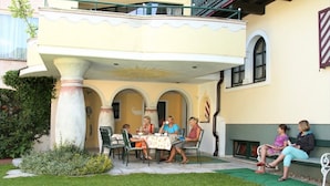 A covered outdoor seating area with a table and chairs, surrounded by a garden and a building with arches and a balcony. - Apartment '12' with Mountain View, Shared Garden and Wi-Fi (Schladming)
