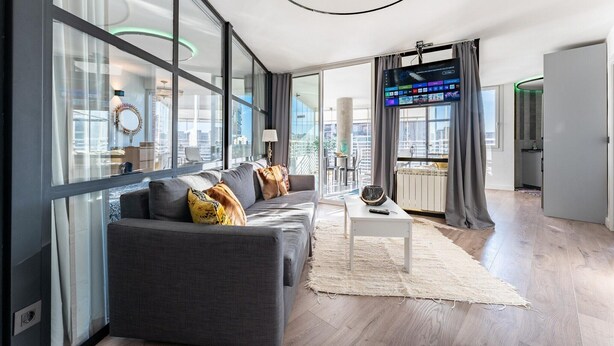 Uma sala de estar moderna com um sofá cinzento, uma mesa de centro branca e uma janela ampla com vista para a cidade.