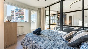 Quarto moderno com uma cama grande, penteadeira de madeira e vista para o horizonte urbano através de amplas janelas.
