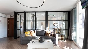 Uma sala de estar moderna com um sofá cinzento, uma mesa de centro branca e uma grande janela com cortinas.