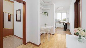 A modern interior with a wooden floor, white walls, a dining area with a table and chairs, a kitchen with a sink and cabinets, and a door leading to another room. - Spacious Apartment | 3 Bedrooms, Balcony, Close to Metro (Warszawa)
