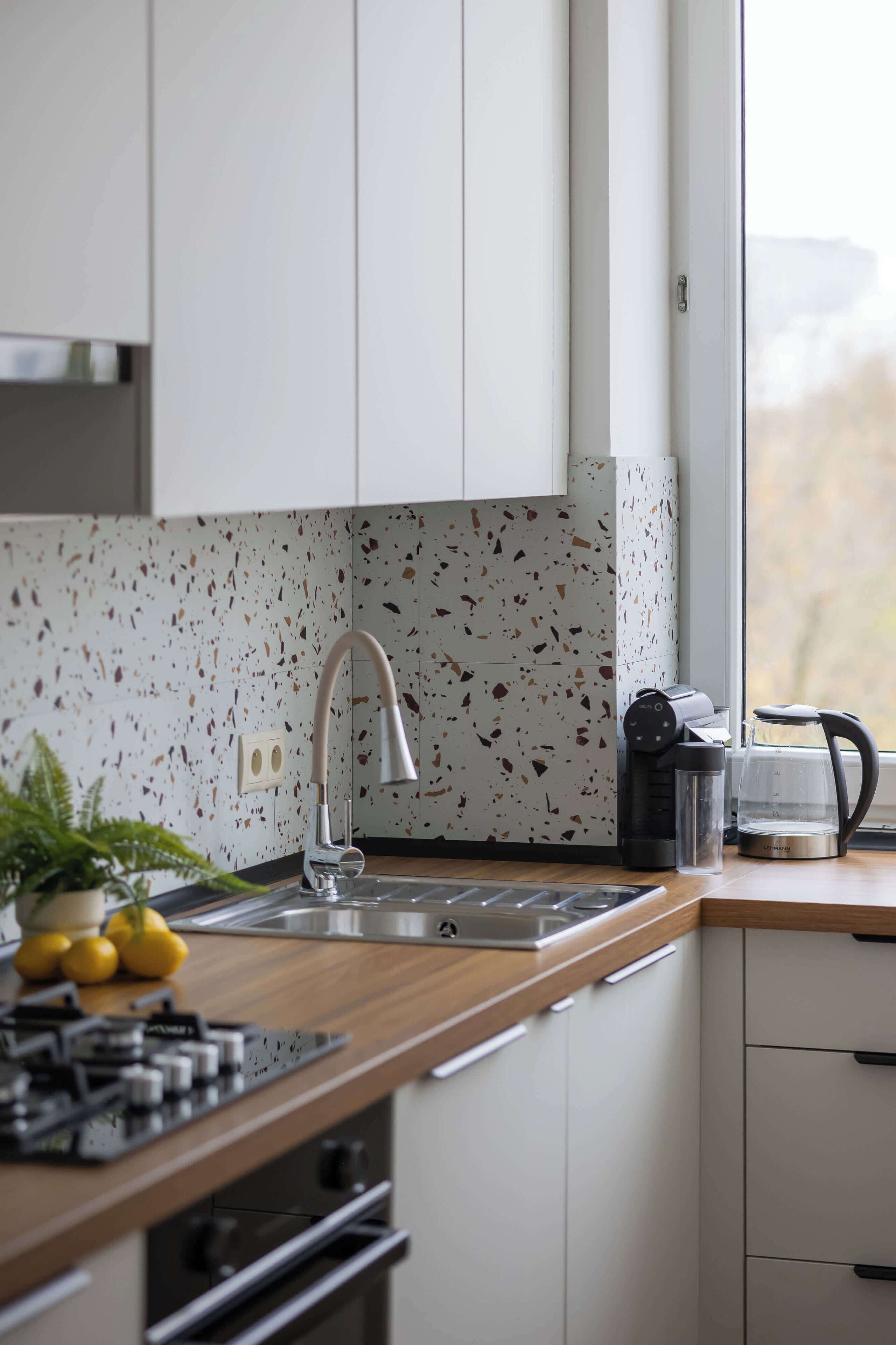 Eine moderne Küche mit weißen Schränken, einer gesprenkelten Spritzschutzleiste, einer Edelstahl-Spüle, einer Kaffeemaschine und einem Fenster mit Aussicht.