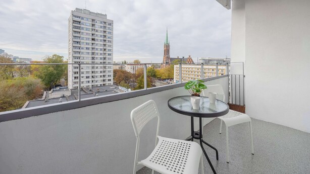 A balcony with a table and two chairs, overlooking a cityscape with buildings and a church spire. - Spacious Apartment | 3 Bedrooms, Balcony, Close to Metro (Warszawa)