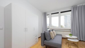A modern room with a grey sofa, a white wardrobe, a small round table, and a window with curtains. - Spacious Apartment | 3 Bedrooms, Balcony, Close to Metro (Warszawa)