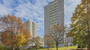 A park with a tall building in the background, benches, and autumn trees. - Spacious Apartment | 3 Bedrooms, Balcony, Close to Metro (Warszawa)