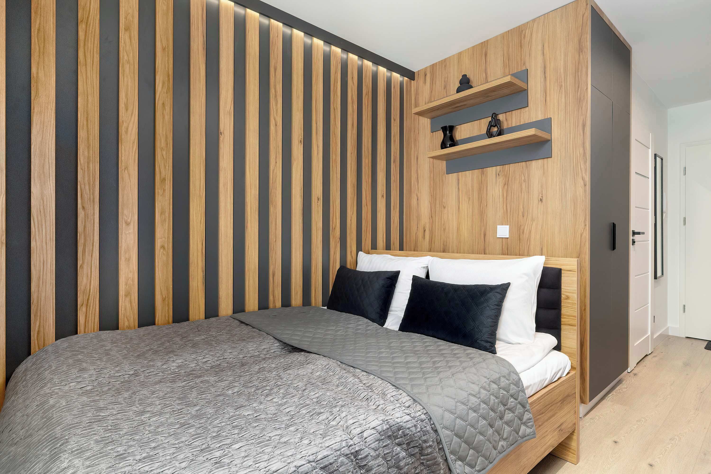 Un dormitorio moderno con cabecera de madera, colcha acolchada gris y dos almohadas negras.