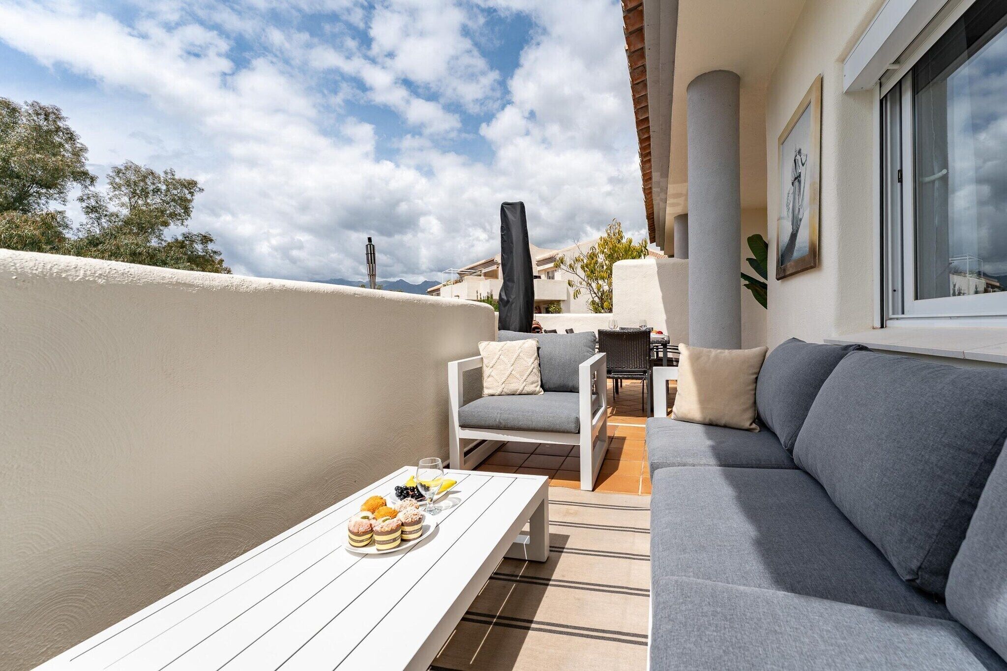 A balcony with a white table set with pastries and drinks, a grey sofa, and a chair with cushions.
