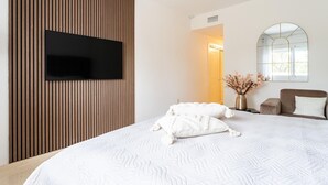A modern hotel room with a large bed, a flat-screen TV mounted on a wooden panel wall, a small seating area with a sofa and armchairs, and a window with a view of the outdoors. - Apartment in Elviria with terrace, Marbella (Malaga)