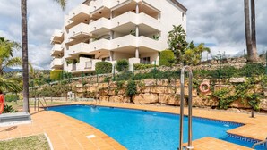 A swimming pool with a tiled deck, surrounded by a building with multiple balconies and a rocky hillside. - Apartment in Elviria with terrace, Marbella (Malaga)