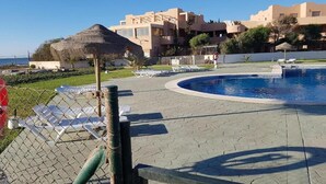 A pool area with lounge chairs, a thatched structure, and buildings in the background.
