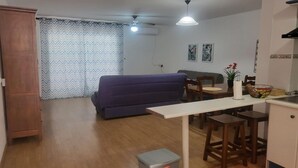 A compact living space with a sofa, a dining table with stools, a small kitchen area, and a ceiling fan.