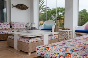 A sunlit room with wicker furniture, a white table, and a view of greenery outside.