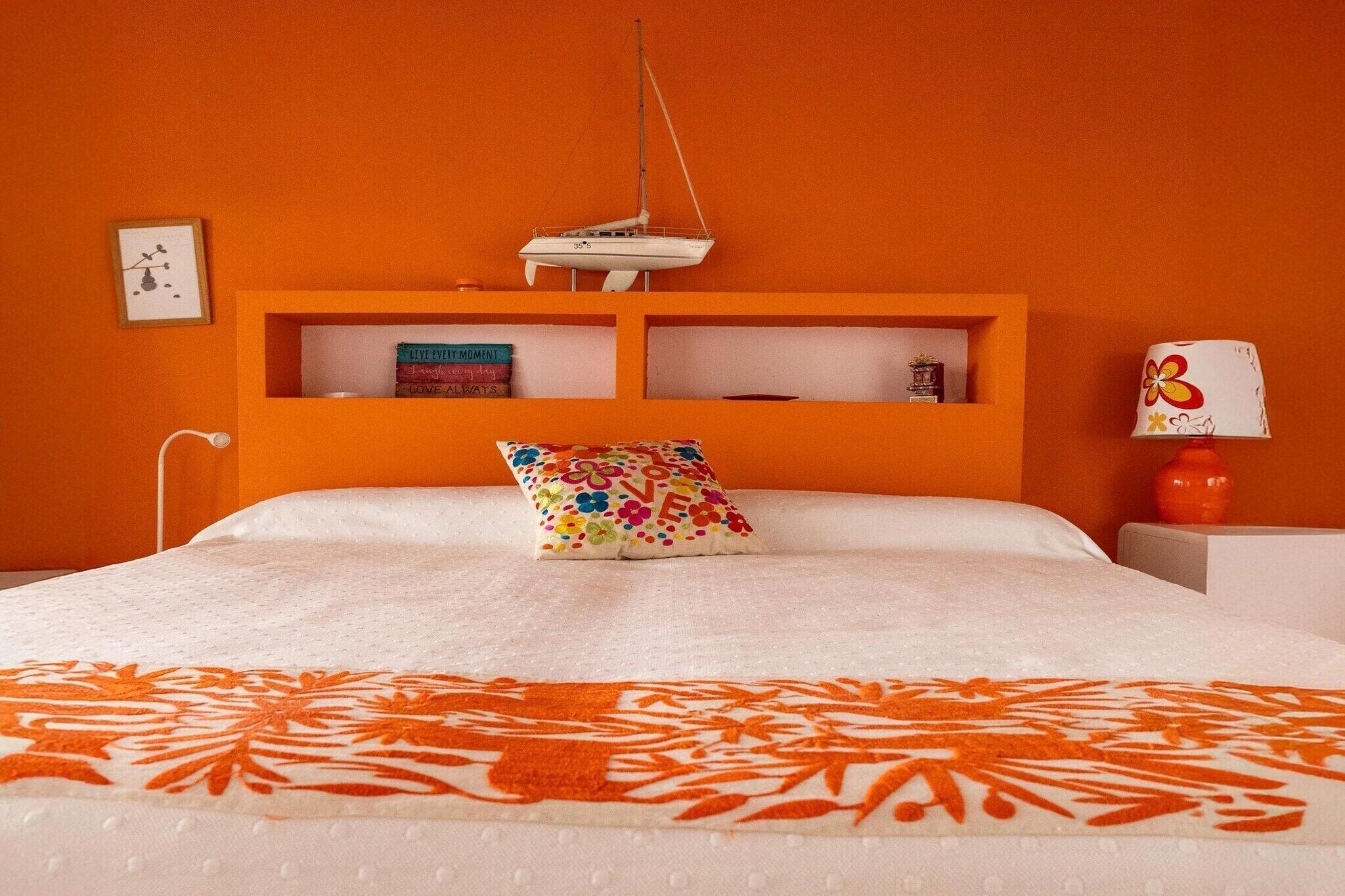 Una cama con un cojín colorido, un pequeño estante con libros y un objeto decorativo en la pared.