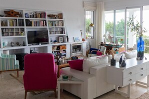 Una sala de estar con un sofá blanco, un sillón rojo, una estantería, un televisor y un ventanal con vistas al follaje.