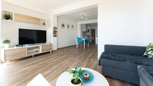 A modern living room with a flat-screen TV, a grey sofa, and a dining area with blue chairs.