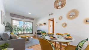 Ein modernes Wohnzimmer mit einem grauen Sofa, gelben Korbstühlen, einem Esstisch mit Obstschale und einem Fernseher im Hintergrund.