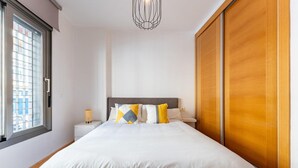 Ein ordentlich bezogenes Bett mit weißer Bettwäsche und Kissen mit geometrischem Muster, eine Nachttischlampe sowie ein Blick durch ein Fenster mit Jalousien.